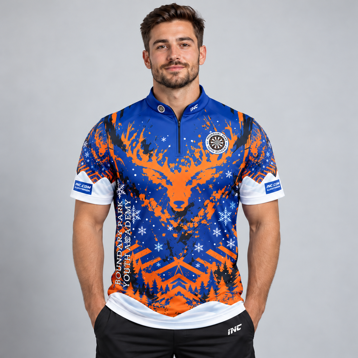 Front view of the TRADE Official Boundary Park Youth Darts Academy Christmas Shirt featuring a festive blue and orange winter scene with reindeer antlers, snowflakes, pine trees, official academy crest, blue zip collar, INC sleeve logos, and Controlled Climate lower hem